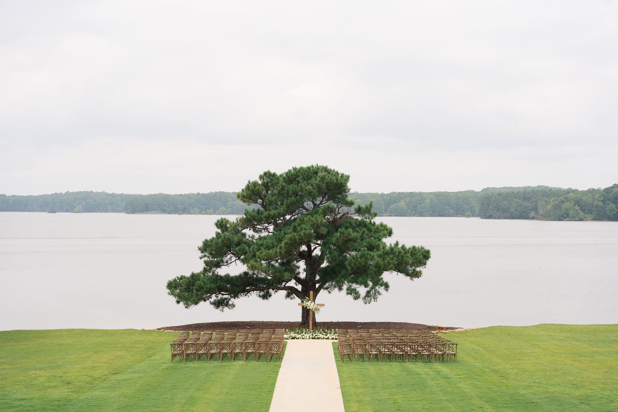 A lush green tree in front of a lake with chairs set up for a wedding ceremony