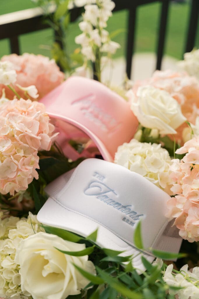 a pink hat along with a white hat surrounded by pink and ivory florals with the names of the newlyweds on their wedding day