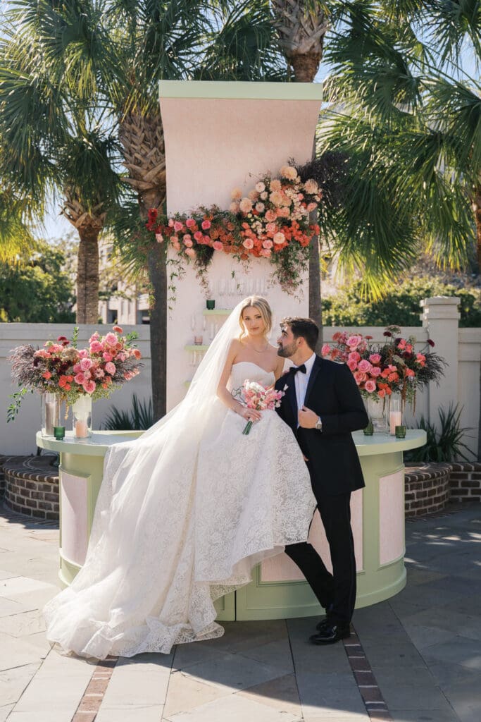 A couple sitting on the bar on their wedding day at the Gadsden House in Downtown Charleston. Surrounded by colorful spring blooms. Photographed by Sara Bartley Photography.