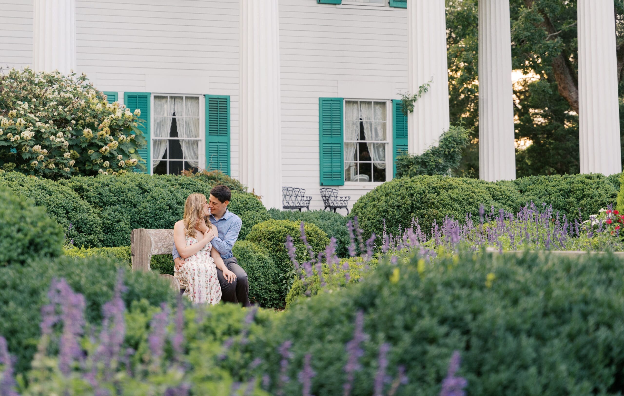 A couple cozied up on a chair surrounded by floral bushes in front of Barrington Hall in Roswell. Photographed by Sara Bartley Photography